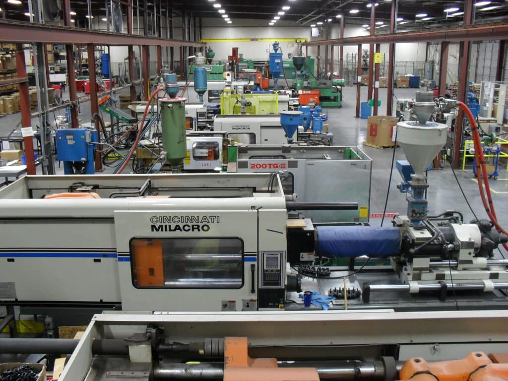 An industrial facility with various plastic injection molding machines, hoses, and material hoppers. The space is organized but complex, with visible infrastructure overhead.