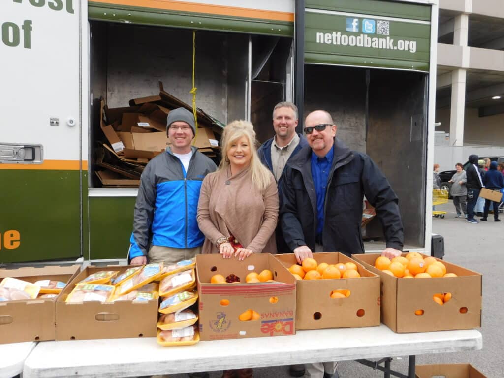 Cuatro personas sonríen delante de un camión de un banco de alimentos con cajas de cartón abiertas llenas de pan y naranjas sobre una mesa.