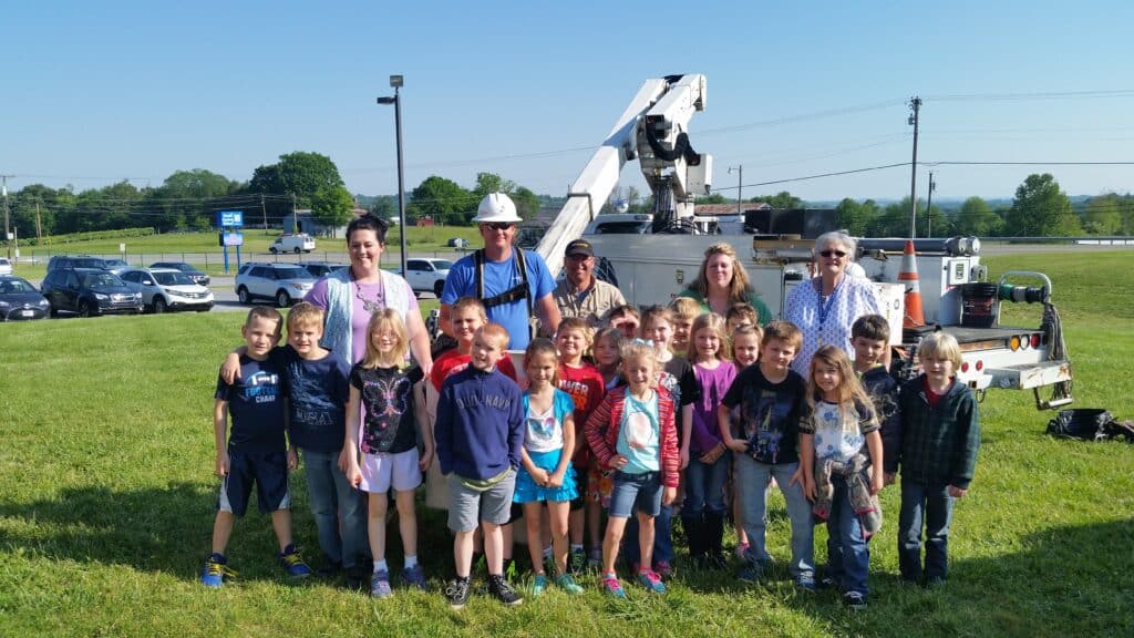 A group of children and adults stand outdoors in front of a utility vehicle with a cherry picker, sunny day, casual clothing, smiling faces.