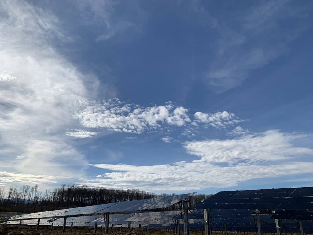 Rows of solar panels under a partly cloudy sky, with trees in the background.