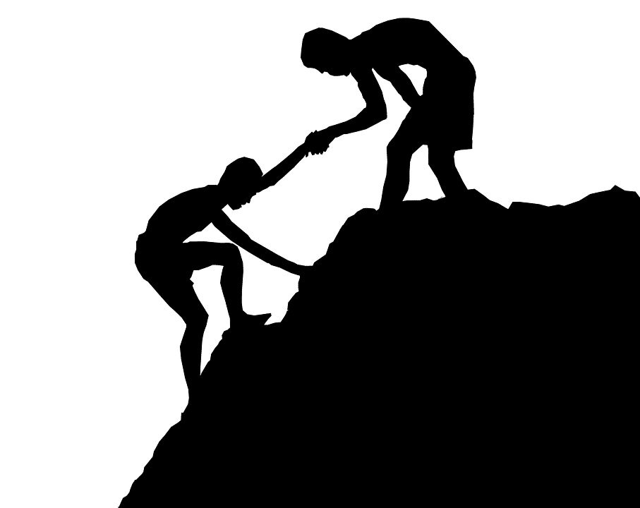 Silhouette of one person helping another climb up a steep rock formation.