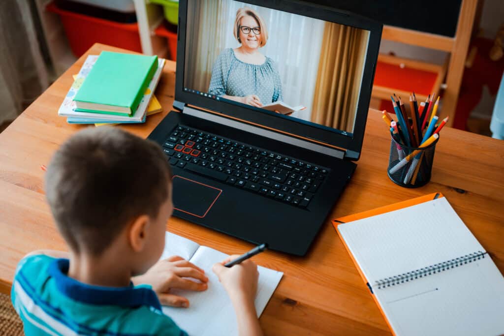 A child takes notes at a desk while participating in a video call with a teacher on a laptop. Nearby are notebooks, a pencil holder, and colored pencils.