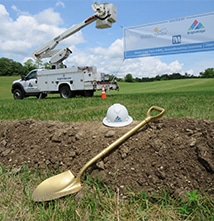 A gold shovel and hard hat in front of a trench, with a utility truck and a banner in the background on a grassy field.