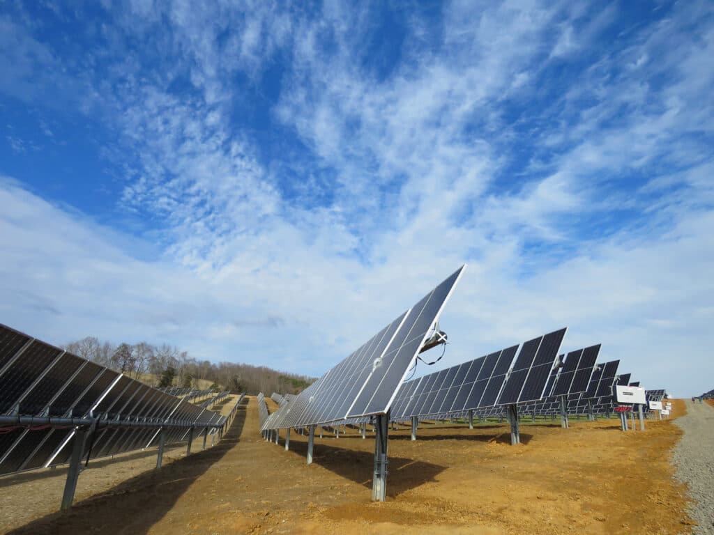 Solar panels arranged in rows on a dirt field under a partially cloudy sky.