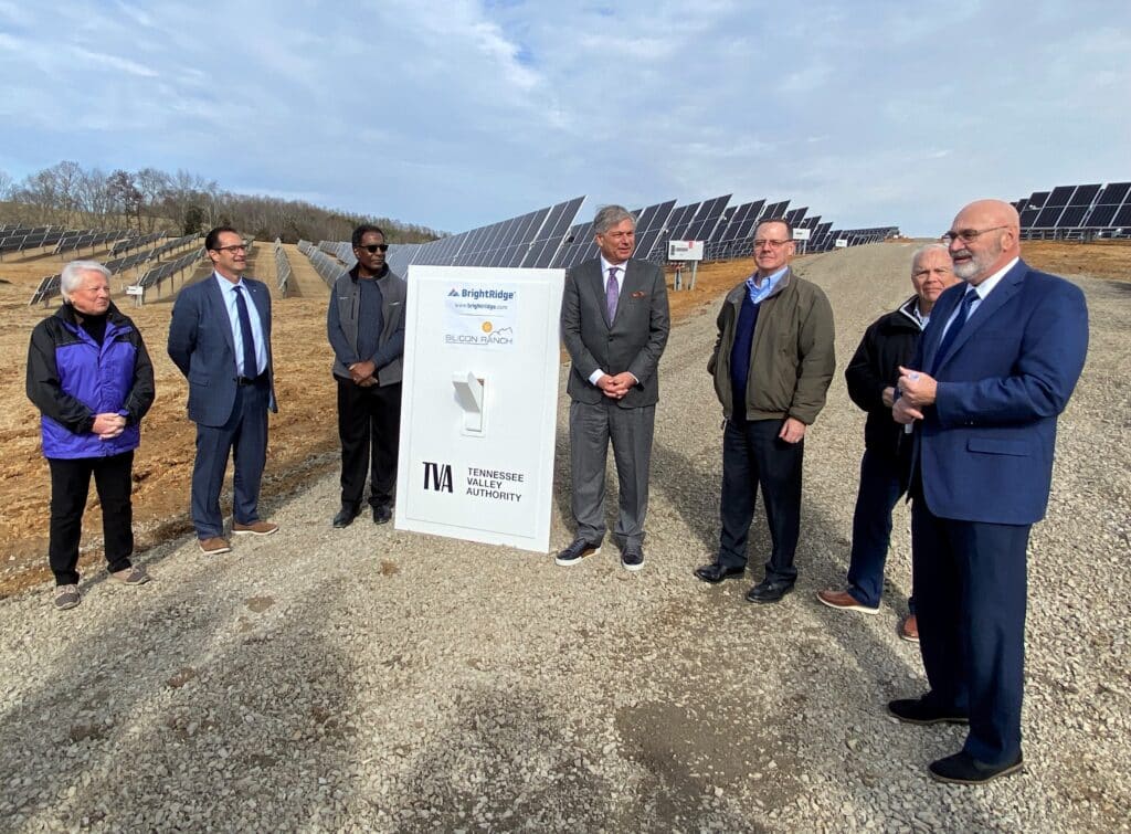 A group of men in suits and jackets stands around a sign in a solar farm, with rows of solar panels visible in the background.