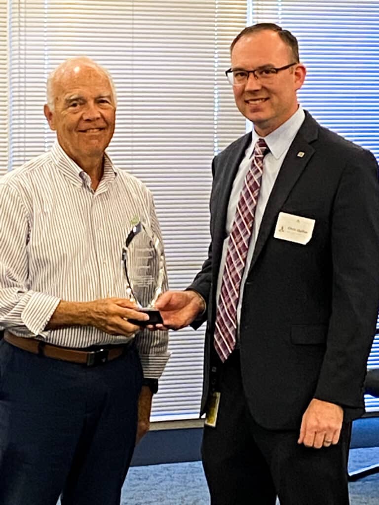 Two men are standing in an office setting. The man on the left, in a striped shirt, is holding an award from TVA EnergyRight, while the man on the right, in a suit and tie, is congratulating him.