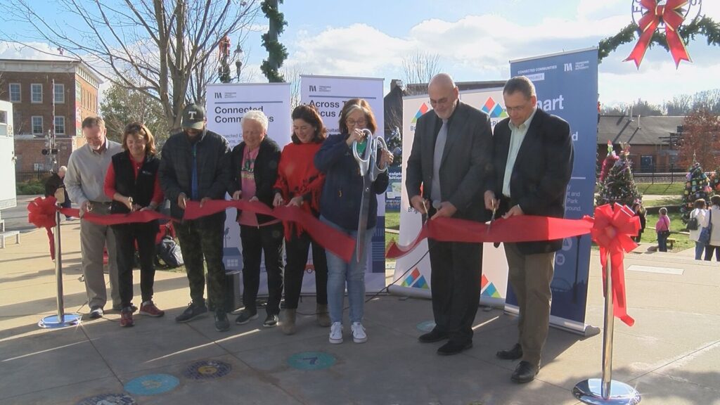 A group of people cut a red ribbon at an outdoor event. Banners in the background display text about community and connectivity initiatives.