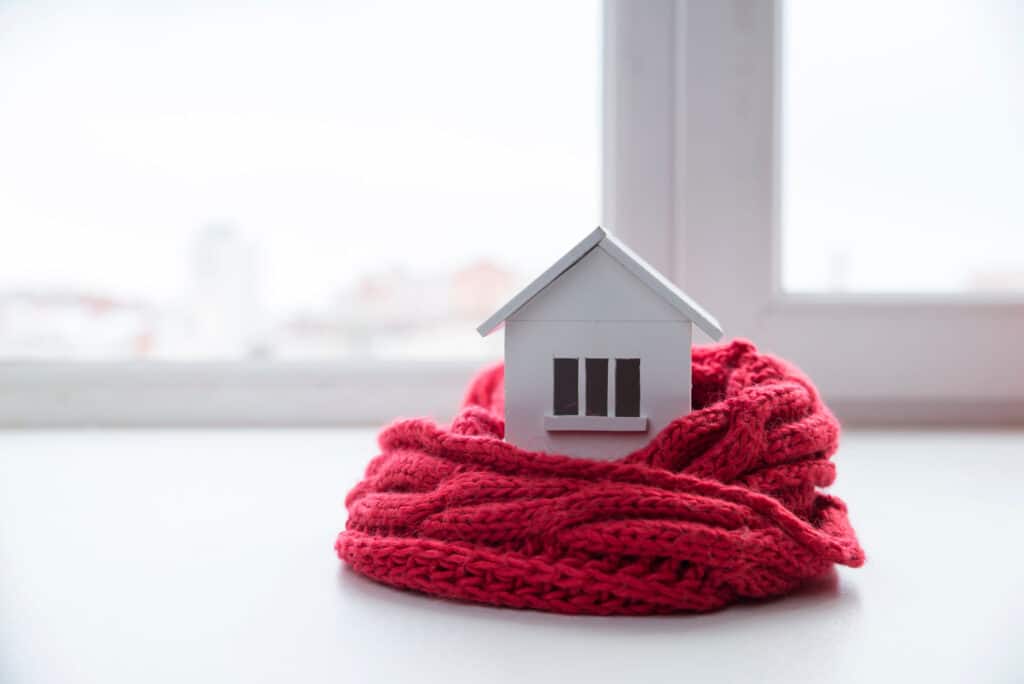 A small white model house wrapped in a red knitted scarf sits on a white surface in front of a window.