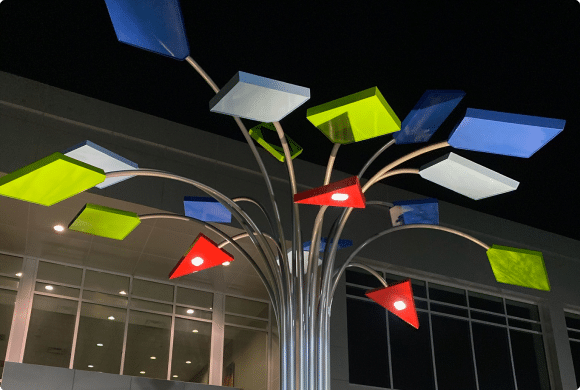 A metal sculpture with colorful geometric shapes, including blue, green, and red, attached to its branches, stands illuminated at night in front of a modern building with large windows.
