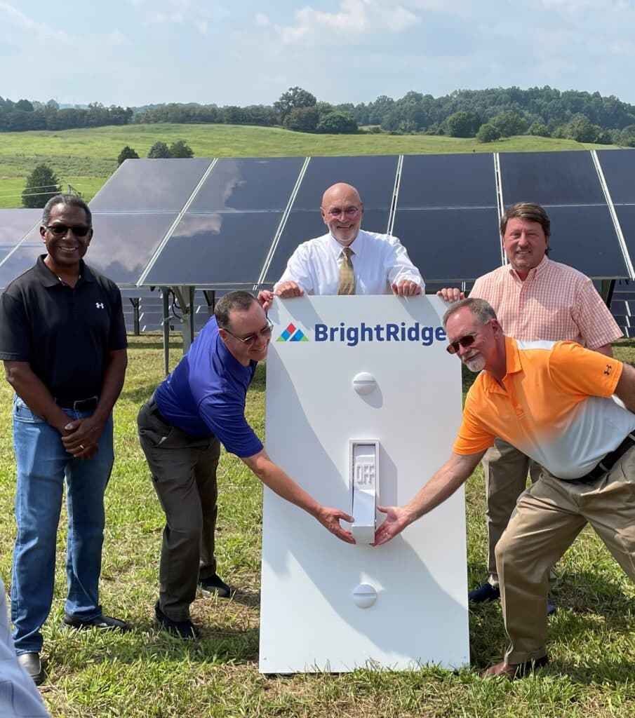 Five men posing around a large model light switch with a BrightRidge logo in front of a solar panel array on a grassy field.