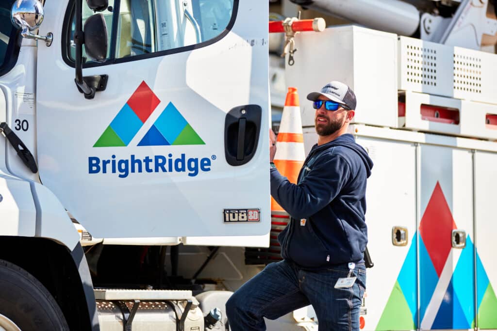 A man with sunglasses and a cap stands next to the open door of a BrightRidge utility truck, holding a cone, with company logos visible on the vehicle.