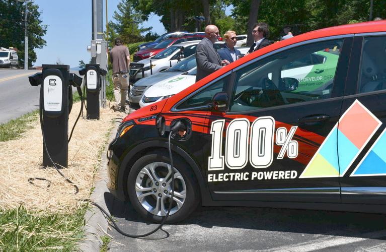 People standing near an electric car charging at a station, with several other vehicles parked and charging in the background. The car has a 100% electric powered label on its side.