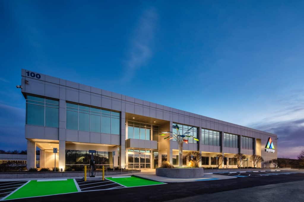 A modern office building with large windows and a colorful sculpture in front. The building is marked with the number "100" and a logo with a colorful triangle. The sky is a gradient of blue.