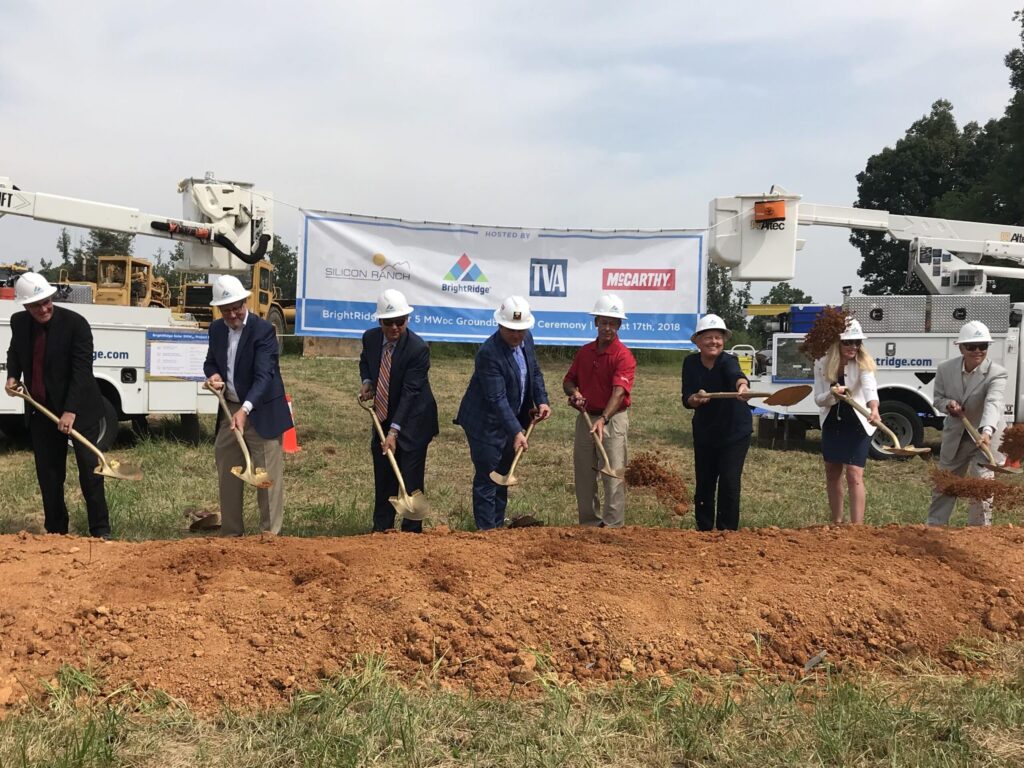 A group of people wearing hard hats and holding shovels participate in a groundbreaking ceremony at a construction site. A banner behind them displays the names of organizations sponsoring the event.